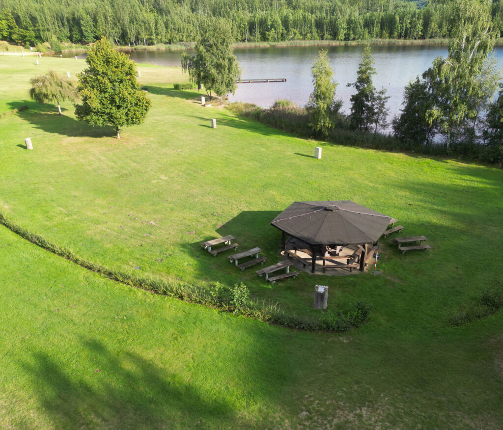 Ein Pavillon aus Holz und Bänke auf einer Wiese am Zieselsmaar-See, umgeben von Bäumen und weitläufigen Grünflächen – ideal für Freizeitaktivitäten oder zum Entspannen in der Nähe des FKK-Bereichs.