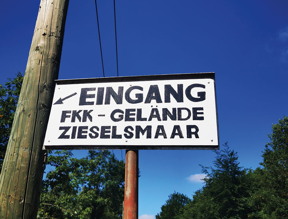 Ein Schild in deutscher Sprache mit der Aufschrift „Eingang FKK-Gelände Zieselsmaar“ lädt Besucher dazu ein, unter blauem Himmel und inmitten grüner Bäume Freizeit und Sport zu genießen.