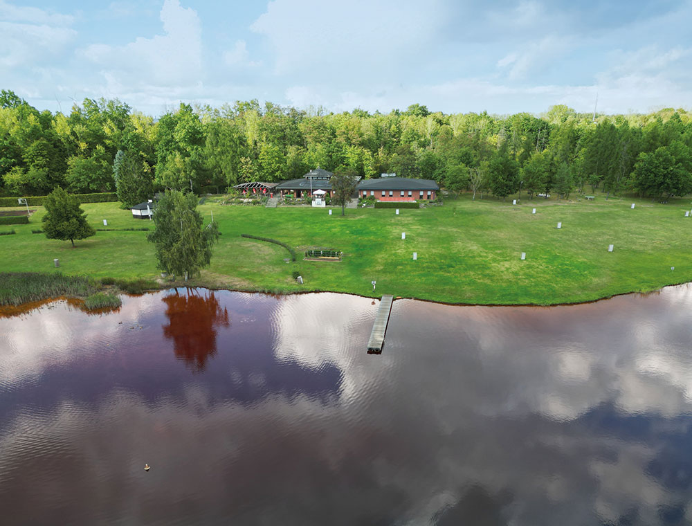 Eine Hütte am Seeufer in Zieselsmaar, umgeben von grünem Gras und Bäumen, mit einem Steg, der in das ruhige, spiegelglatte Wasser hineinragt – ideal für Freizeitaktivitäten oder zum Entspannen in der friedlichen, FKK-freundlichen Atmosphäre.