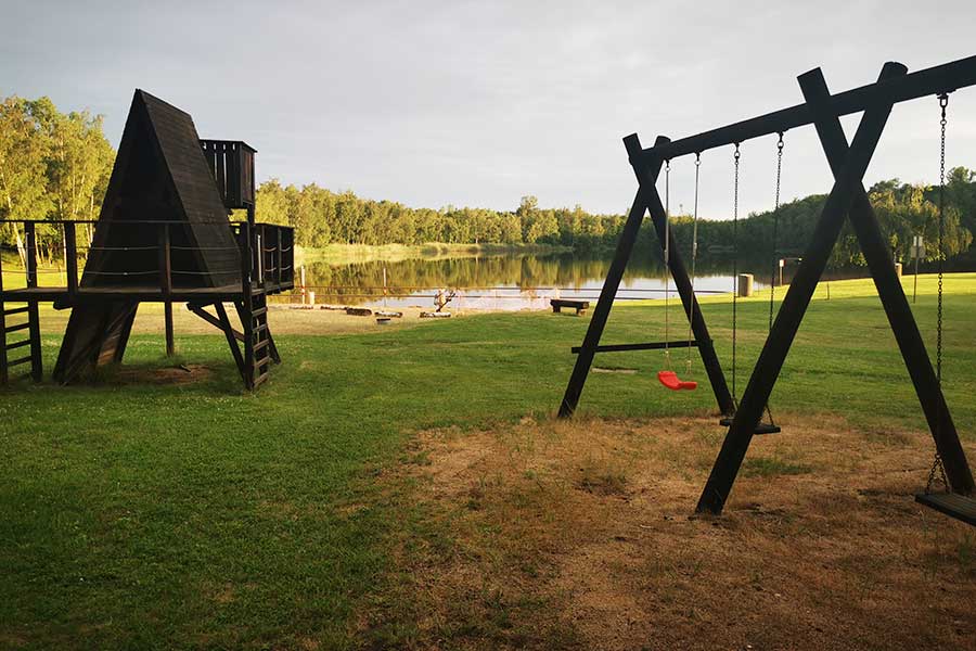 Freizeit-Spielplatz mit Schaukeln und einem Klettergerüst aus Holz in der Nähe eines Sees, umgeben von Rasenflächen und Bäumen.