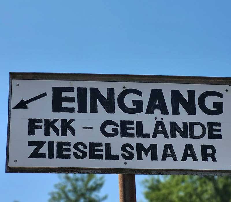 Ein Schild mit der Aufschrift „Eingang FKK-Gelände Zieselsmaar“ und einem Pfeil vor blauem Himmel – es kennzeichnet den Eingang zu diesem beliebten FKK-Sportgelände.