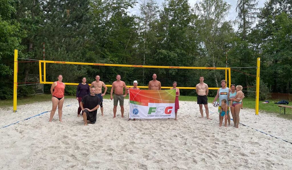 Zehn Personen stehen auf einem sandigen Volleyballfeld in Zieselsmaar und halten ein FSG-Transparent hoch. Die Gruppe genießt Sport und Freizeit, während hohe Bäume eine malerische Kulisse bilden.