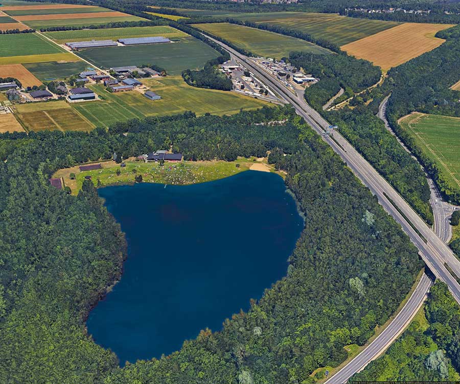 Luftaufnahme eines blauen Sees, umgeben von Bäumen, in der Nähe von Autobahnen und Ackerland – ein idealer Ort für Sport oder zum Entspannen in einem textilfreien FKK-Bereich.