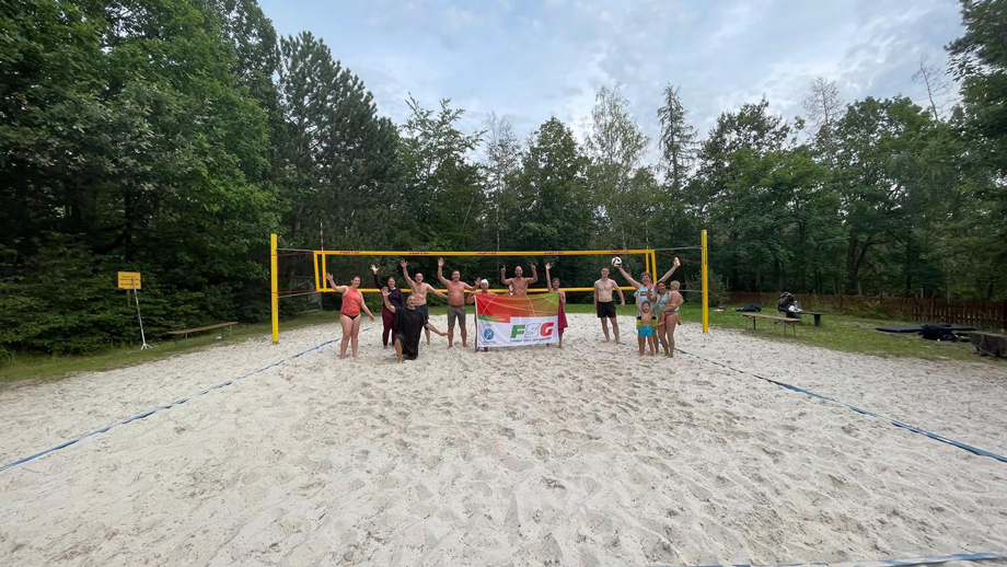 Eine Gruppe von Menschen posiert auf einem sandigen Volleyballfeld, hält ein Transparent hoch und hat Bäume im Hintergrund – gemeinsam feiern sie „Freizeit und Sport“.