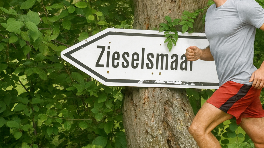 Ein Mann läuft an einem Schild mit der Aufschrift „Zieselsmaar“ vorbei und genießt seine Freizeit vor einem Baum und einer grünen, laubreichen Kulisse.