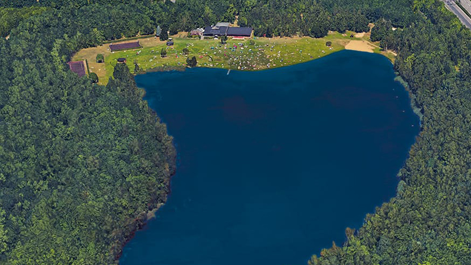 Luftaufnahme von Zieselsmaar, einem herzförmigen blauen See, der von Bäumen umgeben ist, mit einer begrünten Freizeitfläche und Gebäuden am oberen Rand.