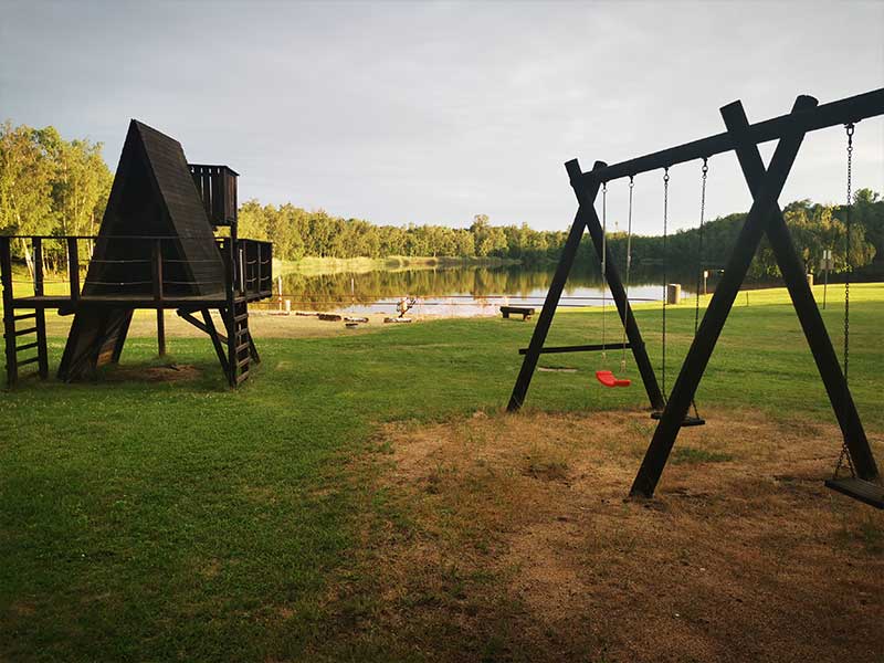 Spielplatz mit Schaukeln und einem Klettergerüst aus Holz in der Nähe des Zieselsmaar-Sees, umgeben von Bäumen – ideal für Freizeitaktivitäten und leichte sportliche Betätigungen.