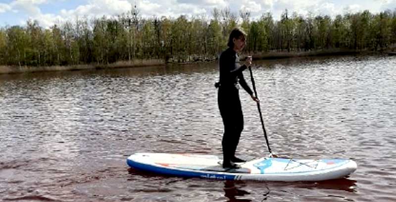 Eine Person in einem Neoprenanzug genießt das Stand-up-Paddleboarding als Freizeitsport auf einem ruhigen See, im Hintergrund von Bäumen umgeben.