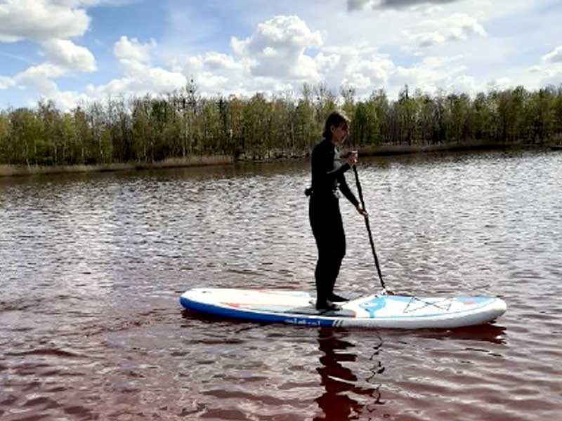 In dieser friedlichen Freizeitszene genießt eine Person das Stand-up-Paddling auf dem ruhigen, rotbraunen Wasser des Zieselsmaar, umgeben von Bäumen und einem bewölkten Himmel.