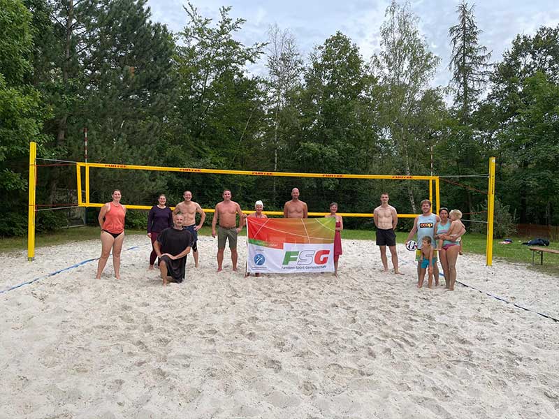 Eine Gruppe von Menschen posiert auf einem sandigen Volleyballfeld, hält ein FSG-Transparent hoch und ist von Bäumen umgeben – sie genießen gemeinsam einen Tag voller Sport und Freizeit.