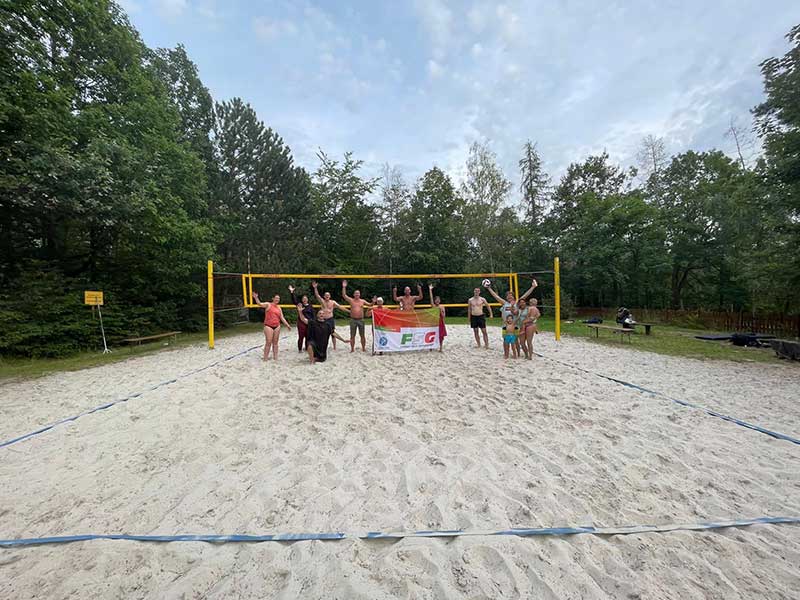 Eine Gruppe von Menschen posiert mit einem Transparent auf dem sandigen Volleyballplatz im Freien in Zieselsmaar und genießt Sport und Freizeit inmitten von Bäumen.