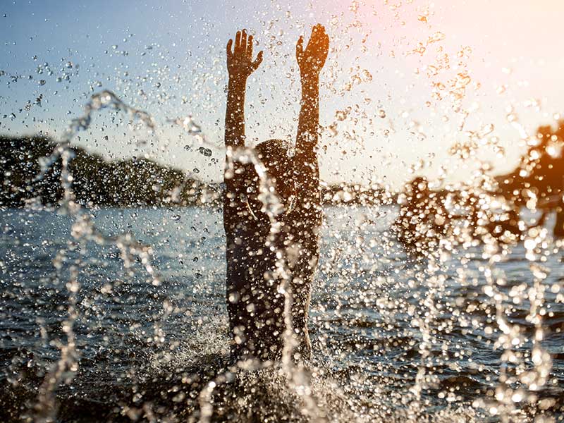 Eine Person, die in Zieselsmaar mit erhobenen Armen im Wasser planscht, während die Sonne scheint und Wassertropfen in der Luft schweben – ein Bild, das die pure Freude an Sport und Freizeit einfängt.